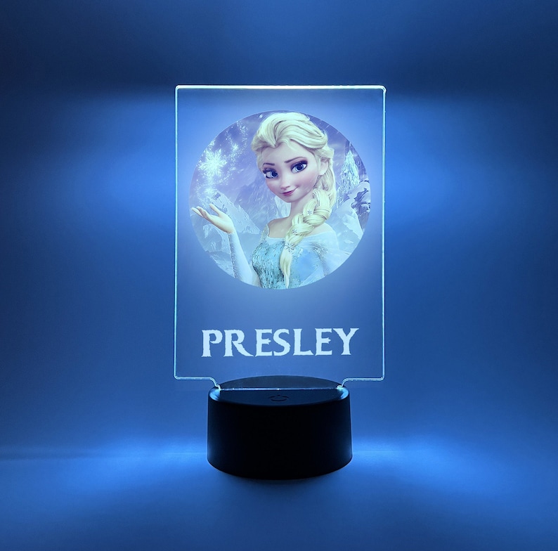 K&ouml;nnte beinhalten: Ein klares Acryl-Nachtlicht mit einem runden Bild von Elsa aus Frozen. Das Bild ist von einem wei&szlig;en Rand umgeben. Der Name "PRESLEY" ist in Wei&szlig; unter dem Bild gedruckt. Das Nachtlicht hat einen schwarzen Sockel mit einem Schalter an der Seite.