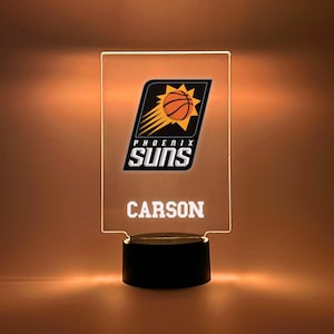 May include: A rectangular LED night light featuring the Phoenix Suns logo with a basketball and sunburst design. The name "CARSON" is displayed below the logo in white lettering. The base is black.