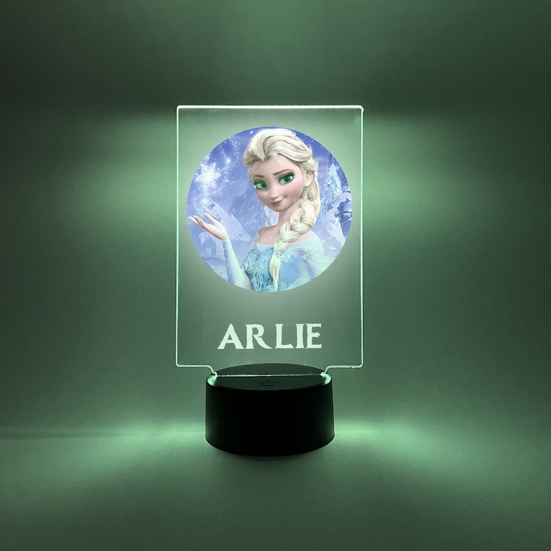 K&ouml;nnte beinhalten: Ein klares Acryl-Nachtlicht mit einem runden Bild von Elsa aus Frozen. Das Bild ist von einem wei&szlig;en Rand umgeben. Der Name "ARLIE" ist in wei&szlig; unter dem Bild gedruckt.