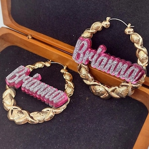 K&ouml;nnte beinhalten: Ein Paar goldene Creolen mit dem Namen "Briana" in pinken und silbernen Glitzerbuchstaben.