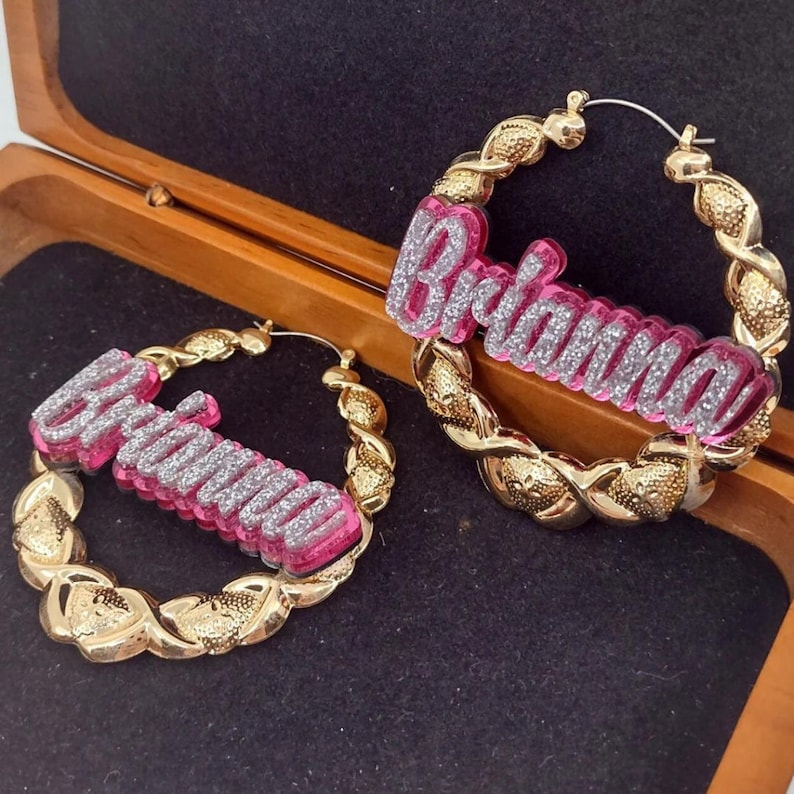 K&ouml;nnte beinhalten: Ein Paar goldene Creolen mit dem Namen "Briana" in pinken und silbernen Glitzerbuchstaben.