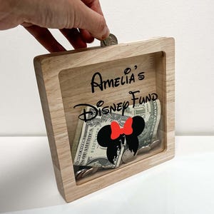 May include: A wooden shadow box with the text "Amelia's Disney Fund" in black script. The box contains dollar bills, coins, and a Minnie Mouse silhouette with a red bow. A hand is dropping a coin into the slot.