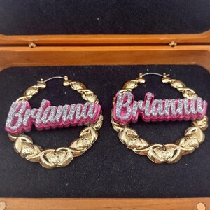 K&ouml;nnte beinhalten: Goldene Creolen mit einem pinkfarbenen und silbernen Glitzer-Namensschild, auf dem "Brianna" steht.