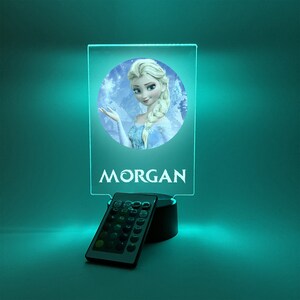 K&ouml;nnte beinhalten: Ein personalisiertes Nachtlicht mit einer durchsichtigen Acrylscheibe, die ein Bild von Elsa aus Frozen zeigt. Die Scheibe wird mit einer Fernbedienung in verschiedenen Farben beleuchtet. Der Name "MORGAN" ist auf die Scheibe gedruckt.