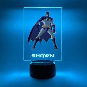 May include: A blue and black Batman character is featured on a clear acrylic night light with the name "SHAWN" printed below. The night light has a black base.