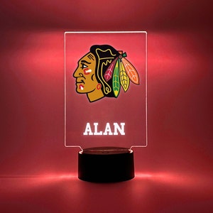 May include: A clear acrylic night light with a black base. The light features a red, black, and green Chicago Blackhawks logo and the name "ALAN" in white text.