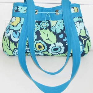 May include: A blue floral tote bag with two long straps. The bag has a drawstring closure and a floral pattern in shades of blue, green, and yellow.