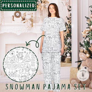 May include: White snowman pajama set with a short-sleeved top and pants. The set features a repeating pattern of snowmen, Christmas trees, and gifts. The name "HUDSON" is incorporated into the design. The words "PERSONALIZED" and "SNOWMAN PAJAMA SET" are also visible.