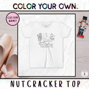 May include: White t-shirt with a black and white outline drawing of a Nutcracker, a ballerina, and a sleigh. The name "Elsie" is written below the drawing. The shirt is ready to be colored in.