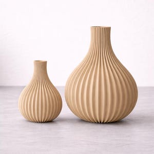 Op de afbeelding: Twee beige, geribbelde vazen van verschillende formaten. De grotere vaas heeft een bolvorm met een smalle hals, terwijl de kleinere vaas een vergelijkbaar ontwerp heeft, maar compacter is. Beide vazen hebben een textuur, verticaal geribbeld patroon.