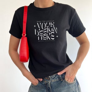 May include: A black baby tee mockup with the text "YOUR DESIGN HERE" and "BABY TEE MOCKUP" and "AESTHETIC MOCKUP STUDIO" printed on the front. The tee is being worn by a person with a red crossbody bag and blue jeans.