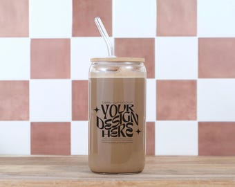 May include: A clear glass cup with a bamboo lid and a curved glass straw, filled with a light brown beverage. The cup has black text that reads "YOUR DESIGN HERE". The background features a checkered pattern in white and dusty rose.