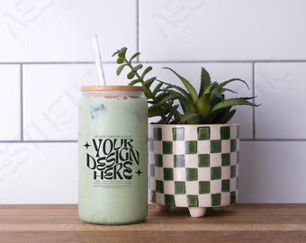 May include: A glass can mockup with a bamboo lid and a straw. The can is filled with a green liquid and has the text "YOUR DESIGN HERE" printed on it. The can is sitting on a wooden surface next to a green and white checkered planter with a green plant.