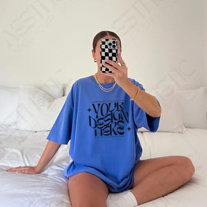 May include: An oversized blue t-shirt with the text "COMFORT COLORS OF YOUR DESIGN HERE" printed on the front. The shirt is being worn by a person sitting on a white bed.