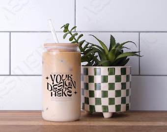 May include: A glass can mockup with a brown lid and a straw. The can is filled with a light brown liquid and has the text "YOUR DESIGN HERE" printed on it. The can is sitting on a wooden surface next to a green and white checkered planter with a green plant.