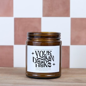 May include: Amber glass candle jar with a white label. The label reads "YOUR DESIGN HERE" in a bold, stylized font. The jar is sitting on a wooden surface against a backdrop of white and terracotta tiles.