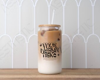 May include: A clear glass can-shaped tumbler with a bamboo lid and a clear straw, filled with a layered drink of iced coffee and milk. The glass has the text "YOUR DESIGN HERE" in black. The tumbler sits on a wooden surface.