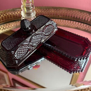 May include: A deep red pressed glass butter dish with a rectangular base and a matching lid. The lid features a textured diamond pattern, circular accents, and a small knob handle. The glass has a translucent finish with intricate detailing.