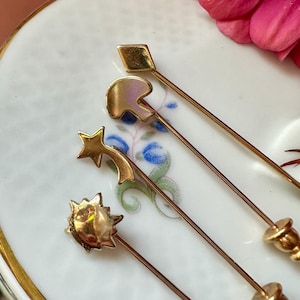 May include: Four gold-toned hat pins with decorative tops. The pins feature a square, a helmet, a shooting star, and a sunburst design. The pins are displayed on a white plate with a floral pattern and a pink flower.