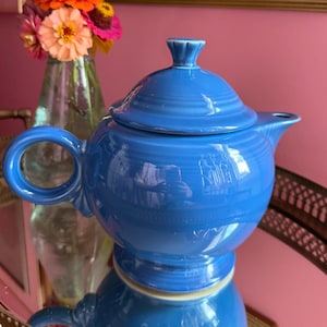 May include: A bright blue ceramic teapot with a matching lid, featuring a rounded body, a circular handle, and a short spout. The teapot has a smooth, glossy finish and a stepped base design, reflecting light on its curved surface.