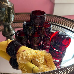 May include: A collection of deep red glass napkin rings featuring a textured diamond pattern on the exterior. One ring holds a yellow fringed linen napkin. The rings are arranged in a stacked formation, showcasing their translucent, dark ruby hue.