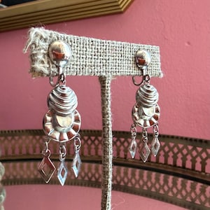 May include: A pair of silver-toned clip-on earrings featuring a smooth round top, a ribbed circular middle section, and a scalloped disc base. Three small diamond-shaped charms hang from the bottom of each earring, creating a tiered design.