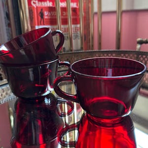 May include: Three ruby red glass teacups with handles are stacked on a mirrored surface. The cups are reflecting the light and the surrounding objects. The cups are part of a vintage tea set.