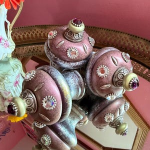May include: A set of three round, mauve-colored decorative finials with a distressed finish. Each finial features silver-toned metal accents, clear faceted crystal inlays, and a small, smooth, light green knob topped with a dark purple gem.