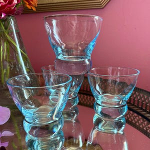 May include: A set of four light blue glass bowls with a wide, flared rim and a thick, rounded base. The glass features a subtle, rippled texture throughout the body, creating a wavy appearance. The bowls are arranged in a cluster on a reflective surface.