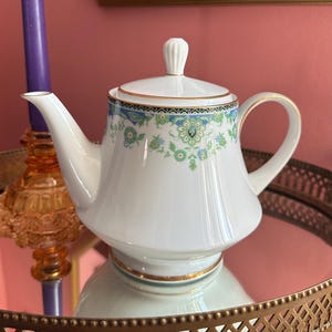 May include: White porcelain teapot with a floral design in shades of blue and green. The teapot has a gold trim and a matching lid with a small handle. The teapot is sitting on a mirrored surface.