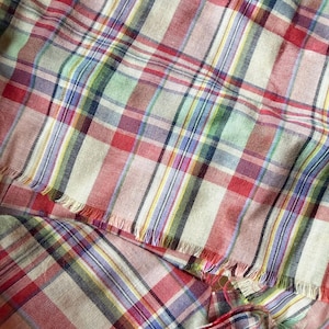 May include: A woven fabric featuring a multi-colored plaid pattern with intersecting lines in shades of red, light blue, green, yellow, and purple on a cream-colored base. The textile has a visible weave texture and frayed edges along the bottom.