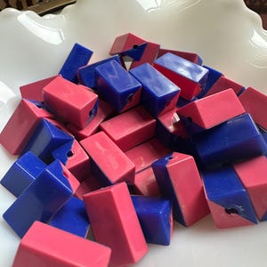 May include: A collection of small, rectangular beads in shades of blue and pink. The beads are piled together in a white, scalloped-edge bowl. Each bead has a hole drilled through it.