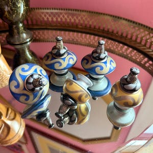 May include: A collection of five decorative ceramic knobs featuring rounded shapes with swirling patterns in blue, gold, and black. Each knob is topped with a small, detailed silver-toned metal finial with intricate engravings and black accents.