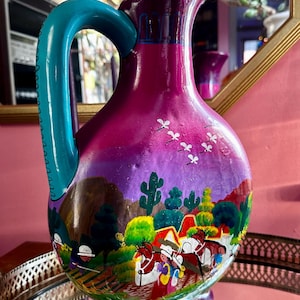 May include: Hand-painted ceramic pitcher with a turquoise handle. The pitcher features a vibrant design with a purple and pink gradient, a desert landscape, and figures on horseback. The decorative jug is a unique piece of art.