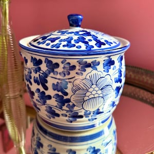 May include: A white ceramic jar with a lid, featuring a blue hand-painted floral pattern. The cylindrical container has a rounded base and a matching lid with a small knob handle, decorated with blue leaves and large flower motifs throughout.
