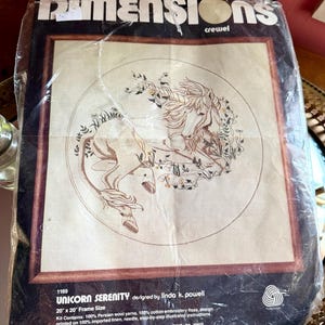 May include: A Dimensions brand crewel embroidery kit titled Unicorn Serenity, designed by Linda K. Powell. The packaging features a line art illustration of a unicorn in a circular floral border, with a 20 inch by 20 inch frame size listed.