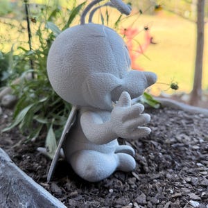 May include: A light gray, cartoonish figurine of a bug-like creature with large eyes, a beak, and antennae. The figure is seated with its hands up, wings extended, and is placed in a planter with dark soil and greenery.