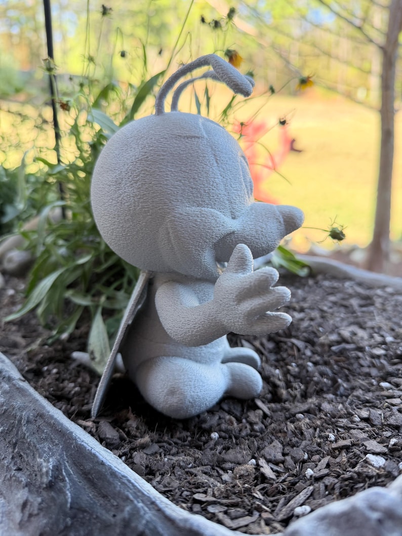 May include: A light gray, cartoonish figurine of a bug-like creature with large eyes, a beak, and antennae. The figure is seated with its hands up, wings extended, and is placed in a planter with dark soil and greenery.