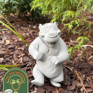May include: A gray concrete statue of a bear with a smiling expression, standing upright. The statue is in a garden setting with mulch and greenery. The logo for Pixie Dust Gardening is in the lower left corner.