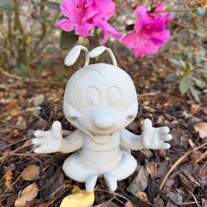 May include: A light gray figurine of a cartoon character with large eyes, antennae, and outstretched arms. The figurine is sitting on a bed of brown mulch, with vibrant pink flowers in the background.