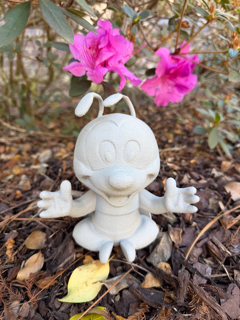 May include: A light gray figurine of a cartoon character with large eyes, antennae, and outstretched arms. The figurine is sitting on a bed of brown mulch, with vibrant pink flowers in the background.