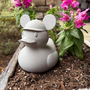 May include: A gray concrete duck figurine with mouse-like ears and a hat, placed in a planter with pink snapdragon flowers. The duck is a whimsical garden ornament, adding a playful touch to the outdoor space.