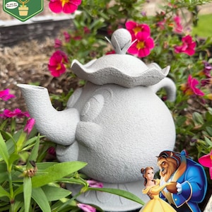 May include: A gray, textured teapot with a floral design, surrounded by pink flowers and green foliage. The teapot has a lid and a spout. The image also includes a cartoon image of Belle and the Beast. The logo "Pixie Dust Gardening" is in the upper left corner.