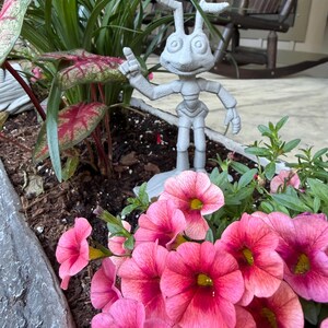 May include: A gray concrete statue of an insect-like character with outstretched arms and a raised finger, placed in a planter box. The planter is filled with pink flowers and green foliage.