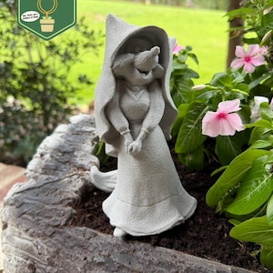 May include: A gray, textured garden statue of a fox character wearing a hooded cloak and a long dress. The fox has a pointed snout and is standing with hands clasped in front. The statue is positioned in dark soil within a rough, bark-like planter.