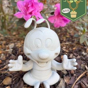 May include: A light gray, cartoon-style garden statue of a character with large eyes, antennae, and outstretched arms. The statue is sitting cross-legged. The background includes pink flowers and a green logo that says "Pixie Dust Gardening".