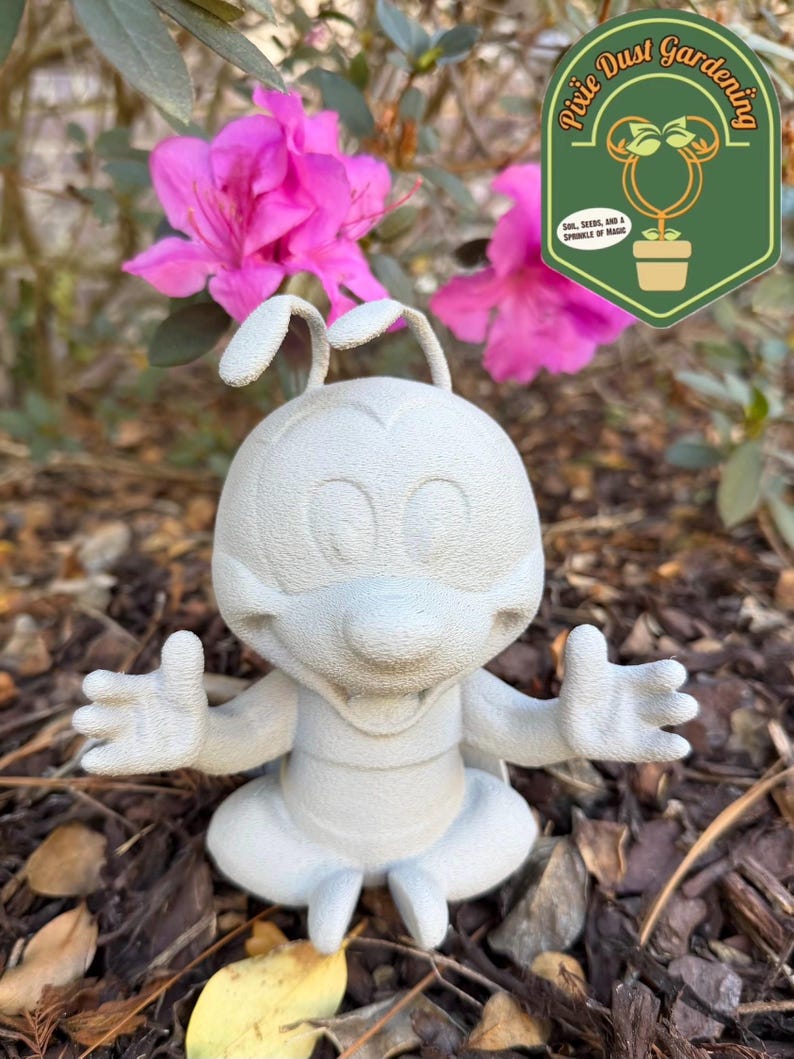 May include: A light gray, cartoon-style garden statue of a character with large eyes, antennae, and outstretched arms. The statue is sitting cross-legged. The background includes pink flowers and a green logo that says "Pixie Dust Gardening".
