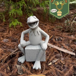 May include: A gray Kermit the Frog statue sitting on a wooden block, with a smiling expression. The statue is set outdoors among brown leaves and green foliage. A logo for Pixie Dust Gardening is in the background.