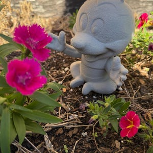 May include: A gray, cartoon-like figurine of an insect with antennae, seated in a garden. The figurine is reaching towards vibrant pink flowers. The flowers have dark green leaves and are in full bloom. The background includes more flowers and a brick wall.