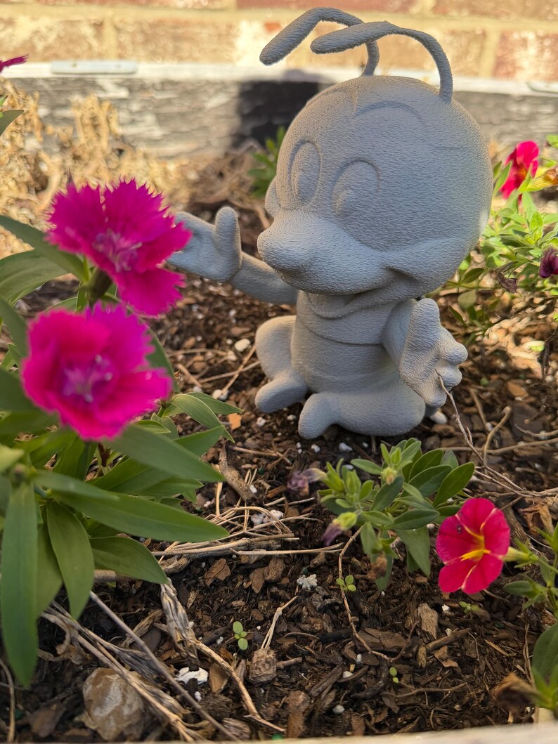 May include: A gray, cartoon-like figurine of an insect with antennae, seated in a garden. The figurine is reaching towards vibrant pink flowers. The flowers have dark green leaves and are in full bloom. The background includes more flowers and a brick wall.